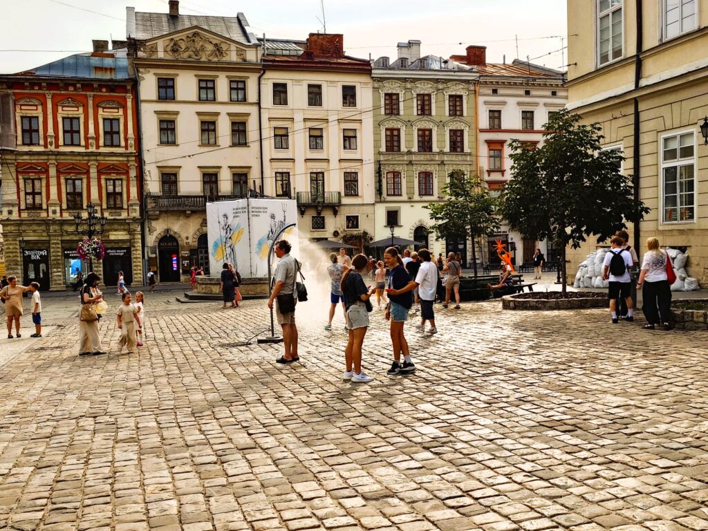 Ein gepflasteter Platz, in der Mitte stehen einige Menschen um einen Duschstrahl. Im Hintergrund weitere Menschen, Bäume, historische Häuser mit bunten Fassaden.