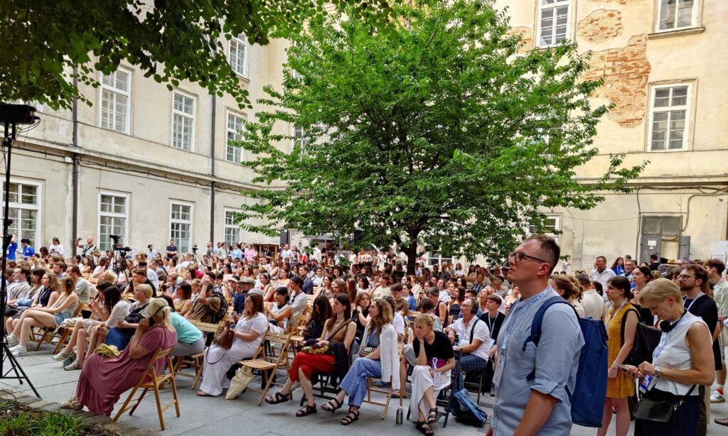 In einem Innnenhof sitzen und stehen zahlreiche Menschen und schauen nach links. Im Hintergrund ein grüner Baum und eine beigefarbene Fassade, stellenweise sichtbares Mauerwerk.