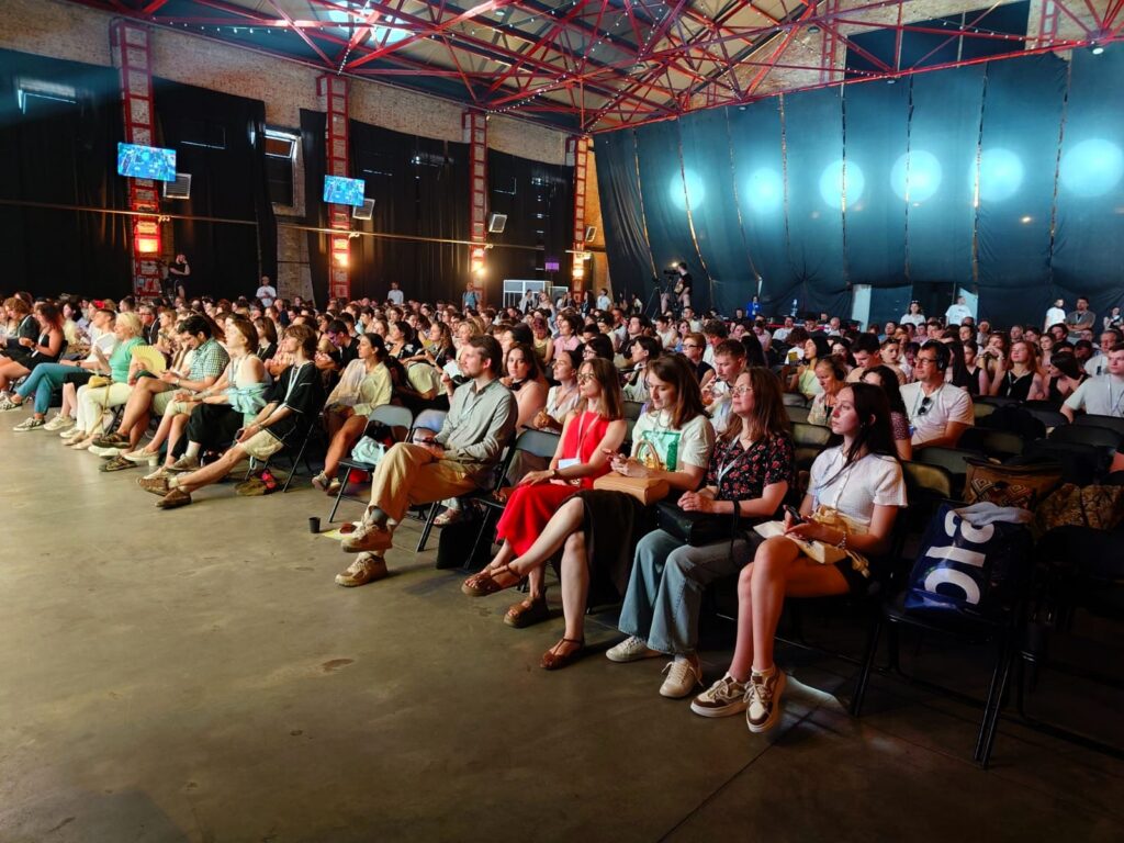 In einer ehemaligen Industriehalle mit Event-Beleuchtung sitzen zahlreiche Menschen auf Stühlen und schauen nach links vorn.