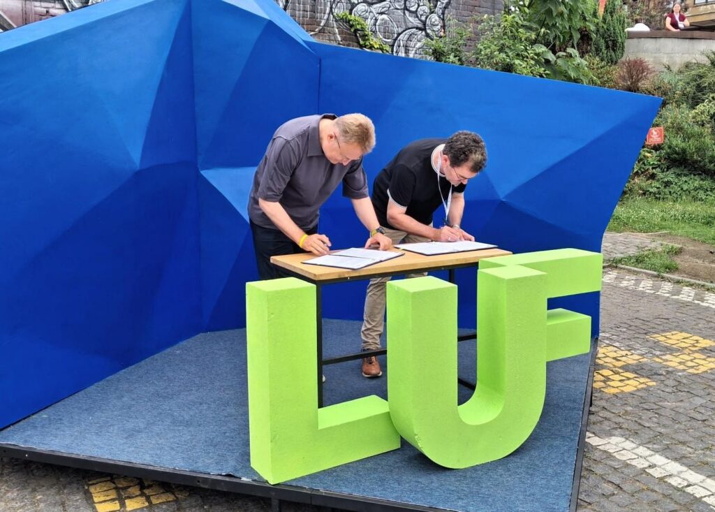 Zwei Männer, der linke mit hellen Haaren, der rechte mit dunklen, beugen sich über einen Tisch und unterschreiben je ein Dokument. Vor ihnen stehen große, grüne Kunststoffbuchstaben. "LUF".