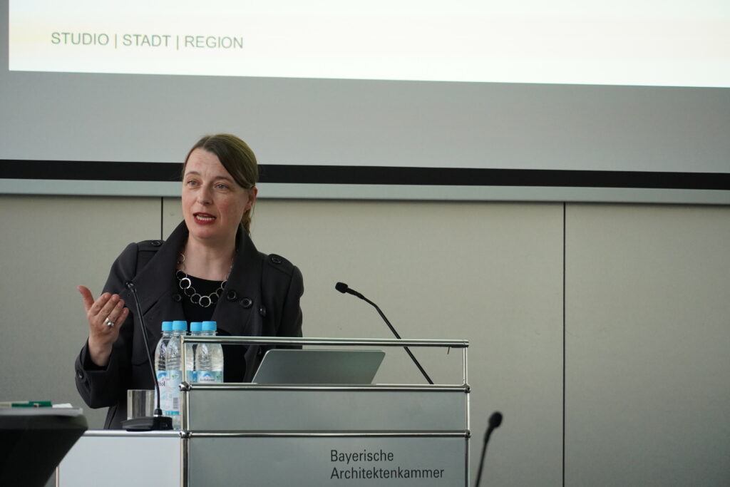 Eine Frau mit hellen Haaren und schwarzer Jacke steht an einem Rednerpult, spricht und gestikuliert.