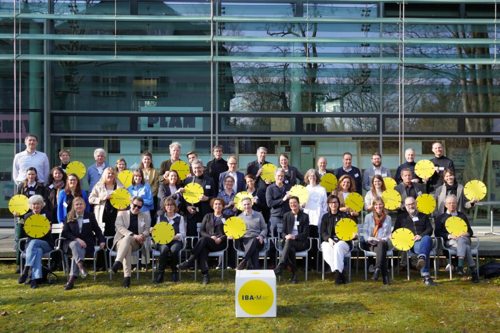 Eine Gruppe von Menschen auf einer Terrasse vor einer gläsernen Fassade. Einige von ihnen halten gelbe Pappkreise.