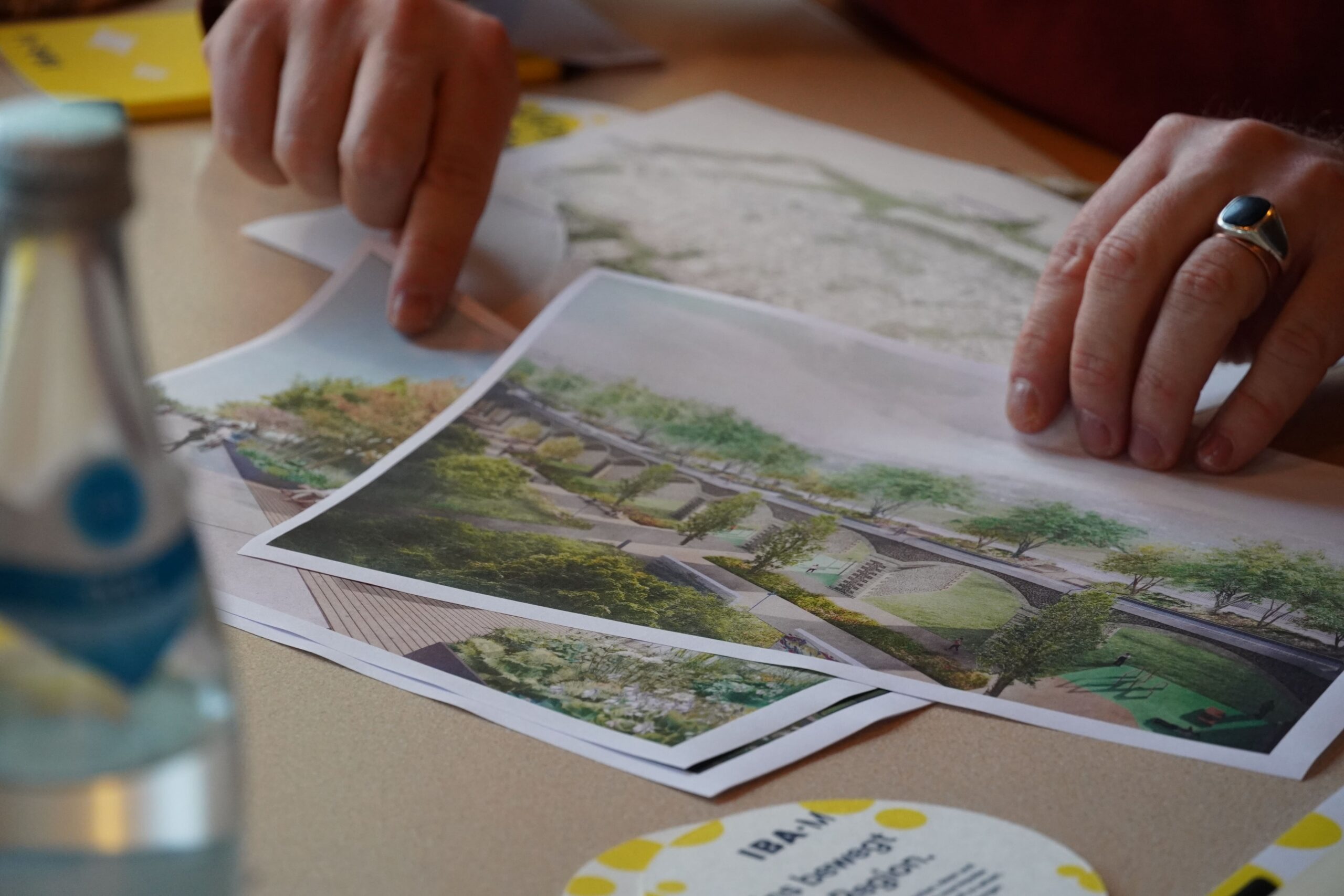 Zwei Hände halten Zeichnungen eines viadukt-ähnlichen Bauwerks, umgeben von einem Park und bepflanzt mit Bäumen.