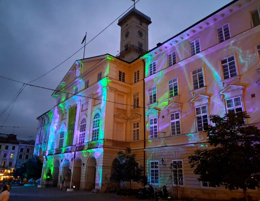 Ein großes, repräsentativ wirkendes Gebäude mit Uhrturm im Dämmerlicht. Auf seiner Fassade sind grüne, türkis- und lilafarbene Lichtstrahlen zu sehen.