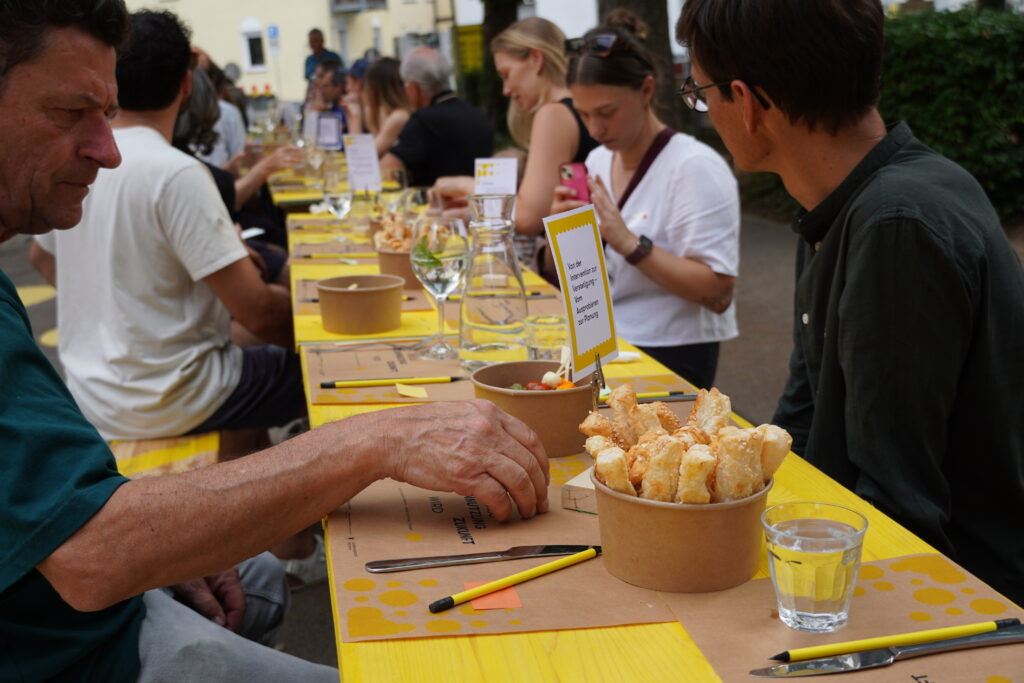 Eine lange gelbe Tafel, rechts und links sitzen Menschen, auf der Tafel Snacks, Gläser, Kärtchen