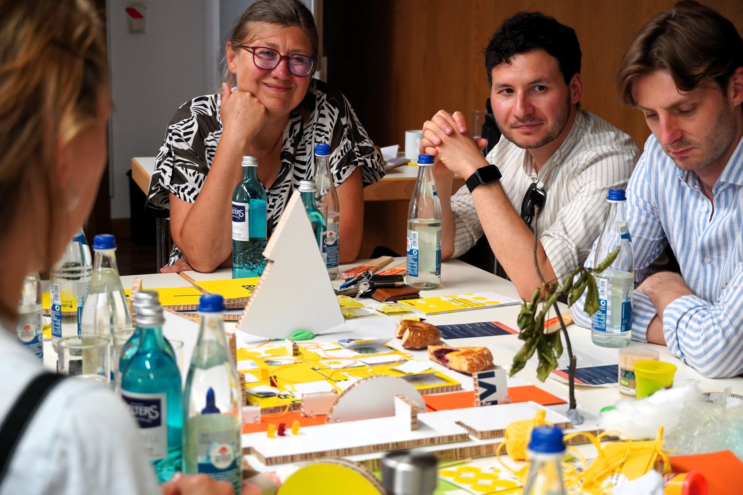 Eine lächelnde Frau und zwei Männer sitzen um einen Tisch, auf dem ein angefangenes Modell aus Pappe, Schnur und Backwaren steht.