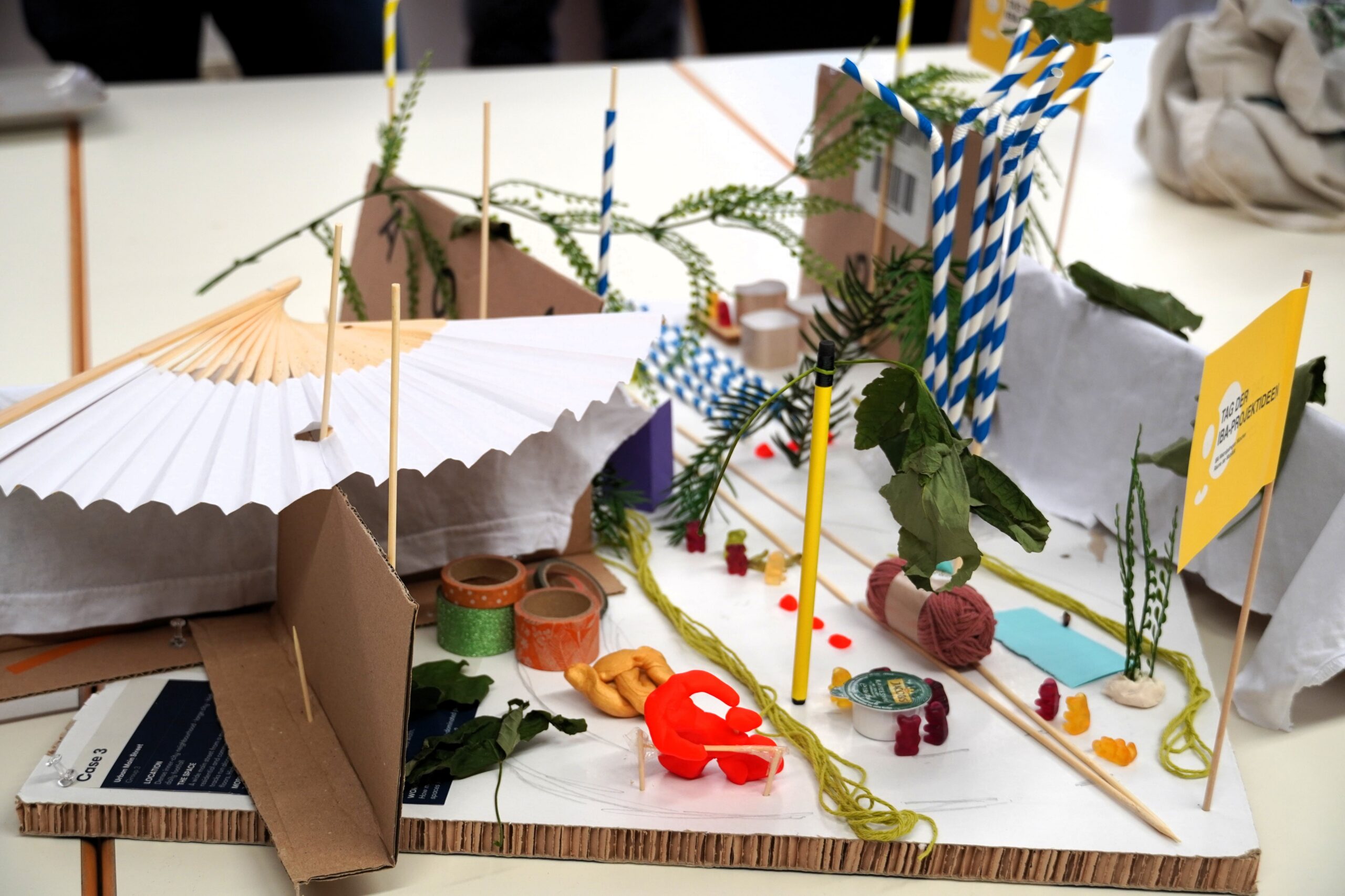 Ein buntes Modell aus Pappe, grünen Zweigen und weiteren Materialien. Links ein Fächer als Überdachung, am Boden Gummibärchen in kleinen Grüppchen, rechts hinten mehrere blau-weiße, aufgestellte Strohhalme.