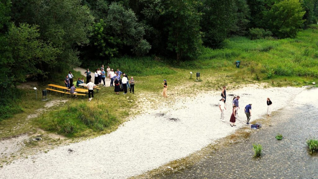 Luftaufnahme eines Flussufers, links unter Bäumen stehen Menschen um Biertische, rechts gehen einige ans Wasser.