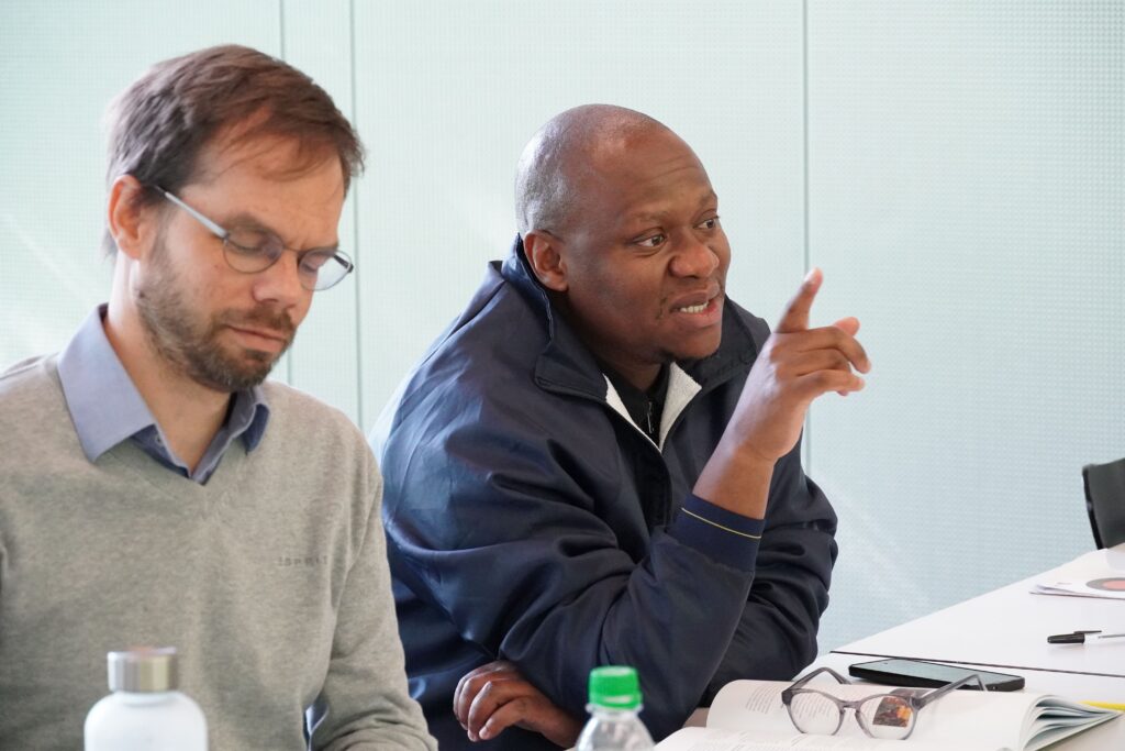Ein Mann in grauem Pullover (links) und einer in blauer Jacke (rechts) sitzen an einem Tisch, der rechte gestikuliert.