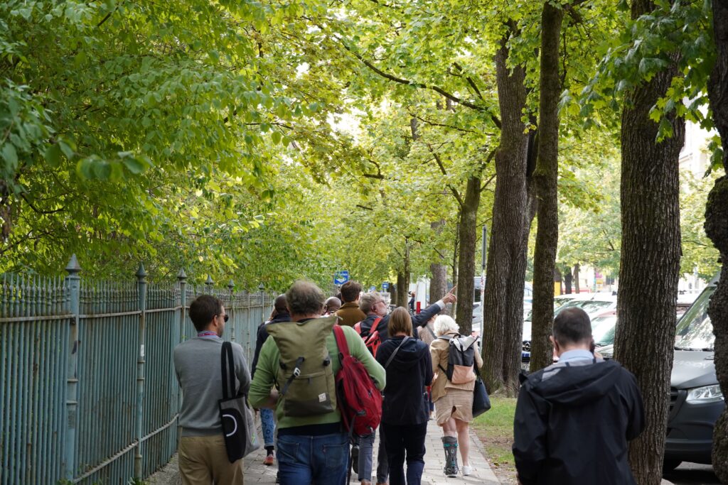 Eine Gruppe von Menschen geht einen Weg entlang, links ein Zaun, rechts Bäume, dahinter parkende Autos.