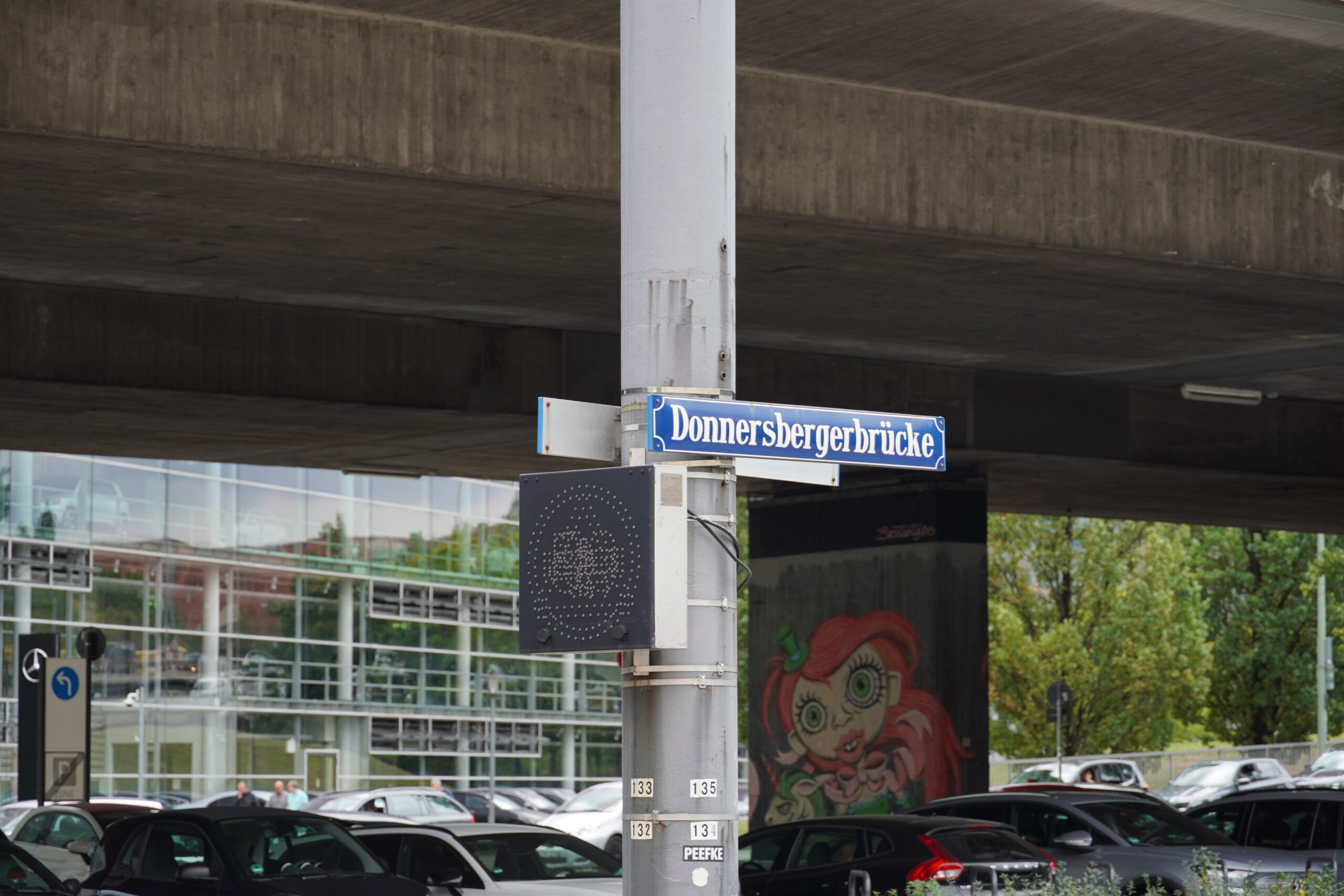 Straßenschild mit der Aufschrift "Donnersbergerbrücke", dahinter schmutziger Beton und parkende Autos.