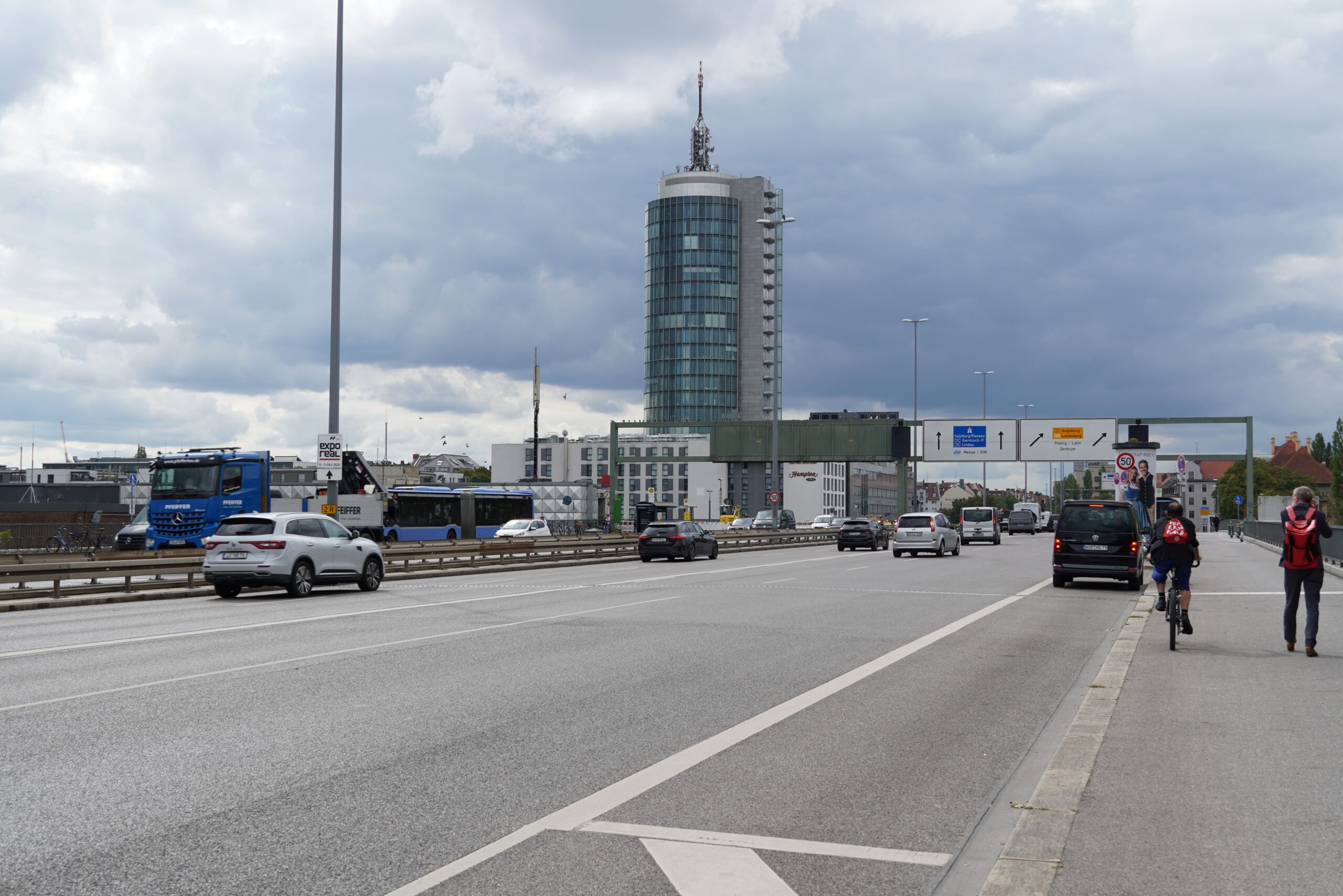 Blick eine Straße entlang, mehrere Autospuren, rechts auf dem Gehsteig eng beieinander ein Radfahrer und ein Fußgänger.