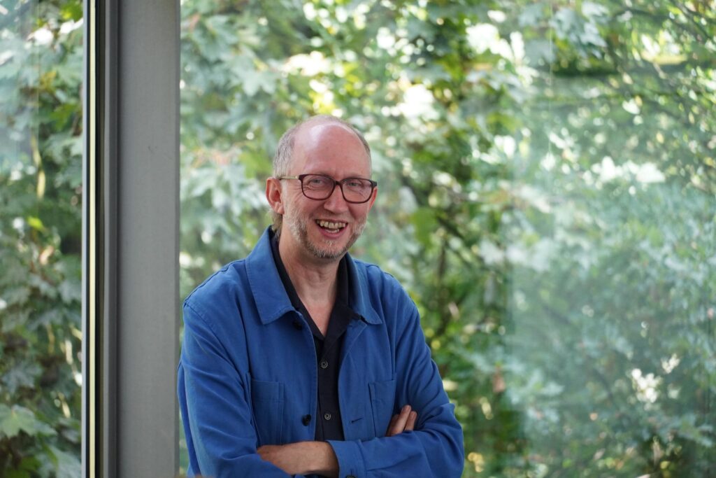 Ein Mann in blauer Jacke verschränkt die Arme und lacht in die Kamera.