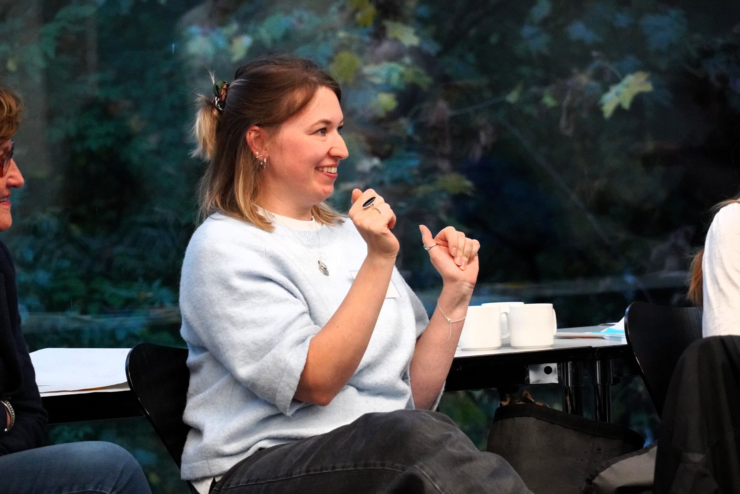 Eine Frau mit hellblauem Pullover, nach rechts gewandt, lächelt und gestikuliert
