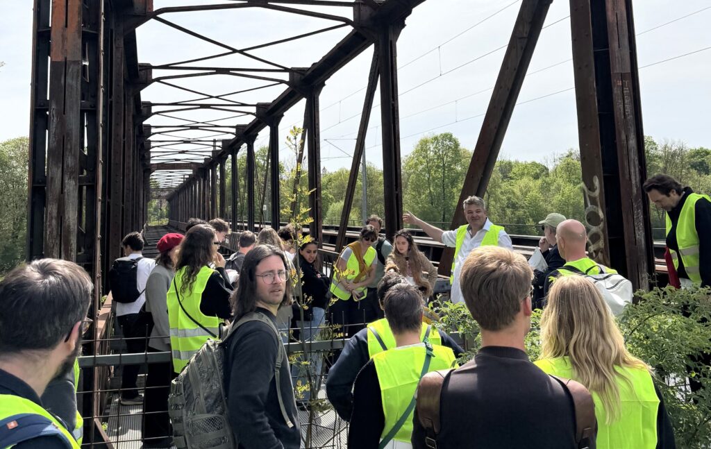 Junge Erwachsene in Warnwesten stehen auf einer Brücke, ein Mann im weißen Hemd deutet ans andere Ende. Über ihnen ragt die rostrote Brückenkonstruktion in den Himmel.