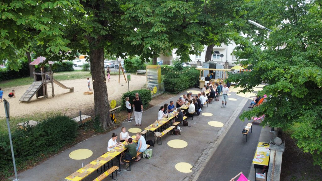 Eine Straße von oben, unter Bäumen steht eine lange Tafel, an der Menschen sitzen